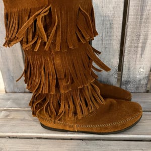 May include: Brown suede boots with multiple layers of fringe detailing. The boots have a moccasin-style sole and a mid-calf height. The color is a warm, earthy brown, and the fringe adds a bohemian flair to the footwear.