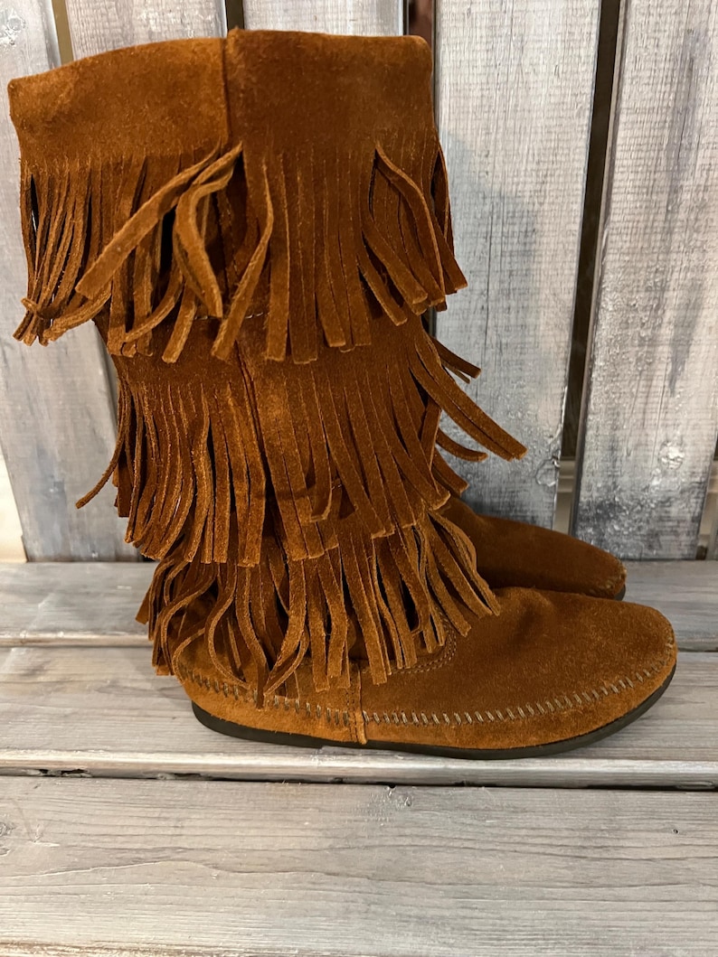 May include: Brown suede boots with multiple layers of fringe detailing. The boots have a moccasin-style sole and a mid-calf height. The color is a warm, earthy brown, and the fringe adds a bohemian flair to the footwear.