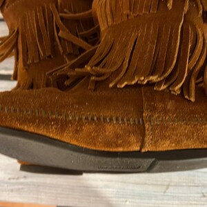 May include: Close-up of brown suede boots with fringe detailing. The boots have a dark brown sole and are made of a soft, textured material. The fringe is long and hangs down from the top of the boot.