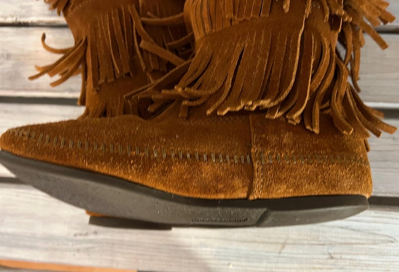 May include: Close-up of brown suede boots with fringe detailing. The boots have a dark brown sole and are made of a soft, textured material. The fringe is long and hangs down from the top of the boot.