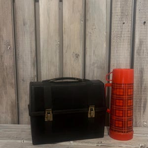 May include: A black lunchbox with a handle and brass-colored clasps stands next to a red plaid thermos. The thermos has a red lid and handle. Both items are on a weathered wooden surface.