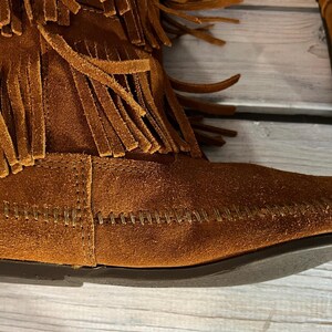May include: Close-up of a brown suede boot with fringe detailing. The boot features a moccasin-style design with visible stitching and a dark sole. The upper part of the boot is adorned with multiple layers of fringe.