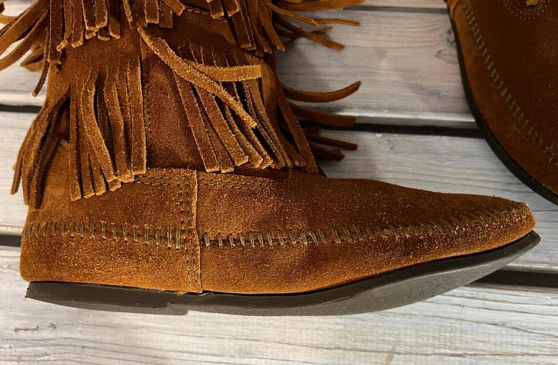 May include: Close-up of a brown suede boot with fringe detailing. The boot features a moccasin-style design with visible stitching and a dark sole. The upper part of the boot is adorned with multiple layers of fringe.