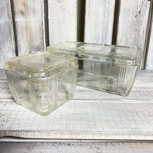 May include: Two clear glass refrigerator storage containers with matching lids. The containers feature a square and rectangular shape with vertical ribbed detailing on the sides and a geometric grid pattern embossed on the top of each lid.