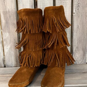 May include: Brown suede boots with multiple layers of fringe detailing. The boots have a moccasin-style toe and a mid-calf height. The boots are made of a soft, suede material and are a warm brown color.