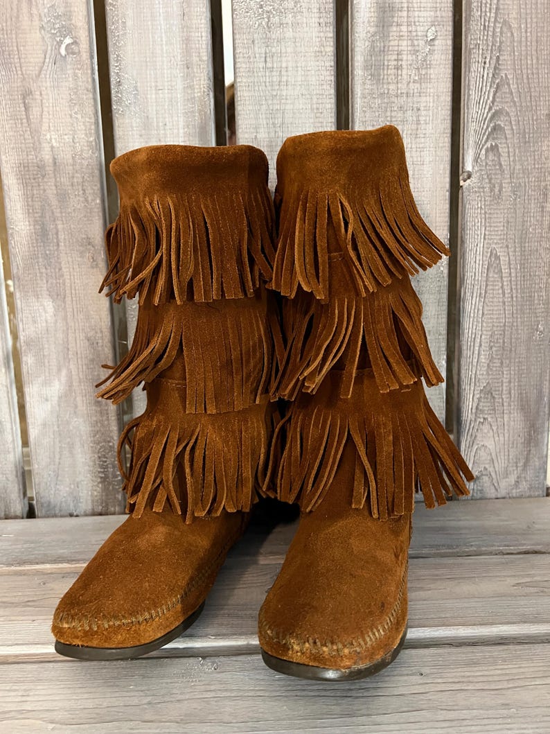 May include: Brown suede boots with multiple layers of fringe detailing. The boots have a moccasin-style toe and a mid-calf height. The boots are made of a soft, suede material and are a warm brown color.
