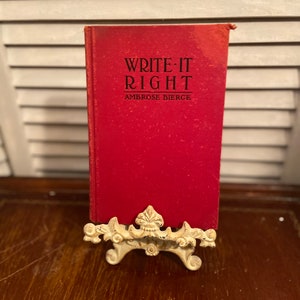 May include: A red hardcover book titled "Write It Right" by Ambrose Bierce, displayed on a white metal book stand.