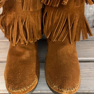 May include: A pair of brown suede boots with multiple layers of fringe detailing. The boots have a moccasin-style construction with visible stitching around the soles. The boots are a warm, earthy tone, suggesting a casual or bohemian aesthetic.
