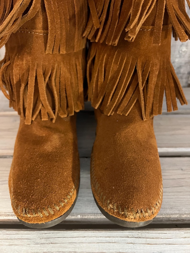 May include: A pair of brown suede boots with multiple layers of fringe detailing. The boots have a moccasin-style construction with visible stitching around the soles. The boots are a warm, earthy tone, suggesting a casual or bohemian aesthetic.