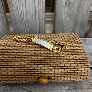 May include: A gold-toned bracelet with a rectangular, mother-of-pearl center piece rests on a woven bamboo box. The bracelet features a chain link design. The box has a wooden knob closure and is made of light brown bamboo.