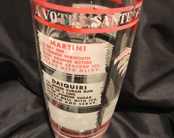 Op de afbeelding: Een glazen cocktailshaker met rode en witte strepen en de tekst "A Votre Santé" erop gedrukt. De shaker bevat ook instructies voor het maken van een Martini en een Daiquiri.