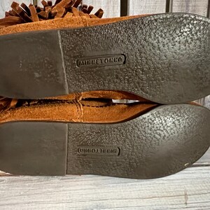 May include: Close-up of the soles of two brown suede moccasin boots. The soles are dark brown with a textured pattern and the brand name "MINNETONKA" is embossed on each sole. The boots have a fringe detail.