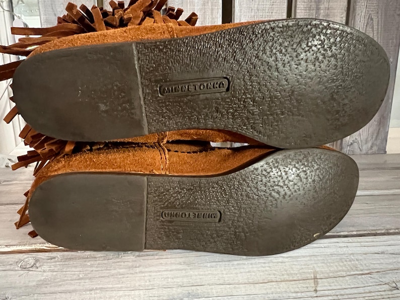 May include: Close-up of the soles of two brown suede moccasin boots. The soles are dark brown with a textured pattern and the brand name "MINNETONKA" is embossed on each sole. The boots have a fringe detail.