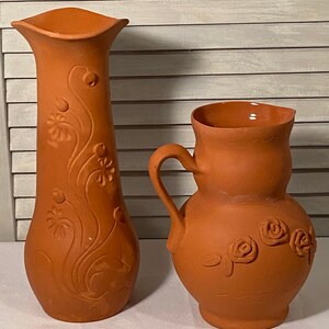 May include: Two terracotta pottery pieces, a tall vase with a floral design and a shorter pitcher with a rose design.