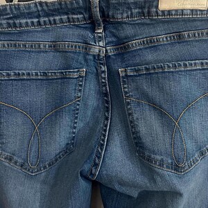 May include: Close-up of the back of blue denim jeans. The jeans have two back pockets with a curved stitch design. The Calvin Klein Jeans label is visible at the waistband. The jeans are a medium wash.