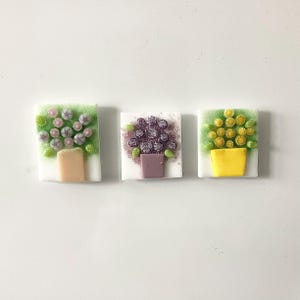 May include: Three square glass art pieces depicting floral arrangements in pots. The first features pink and purple flowers with green leaves and a tan pot. The second has purple flowers and a purple pot. The third has yellow flowers and a yellow pot.
