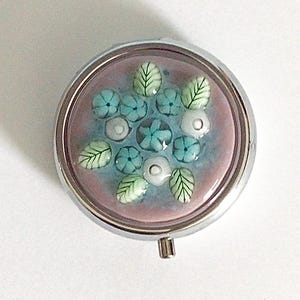 May include: A round, silver-toned pill box with a floral design. The interior features a pink and blue background with turquoise flowers, white accents, and green leaves. The pill box has a hinged lid and a small clasp.