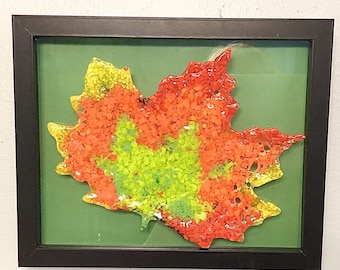 May include: A framed artwork featuring a colorful leaf design. The leaf is composed of small, textured pieces in shades of yellow, orange, and green, set against a green background. The artwork is encased in a black frame.