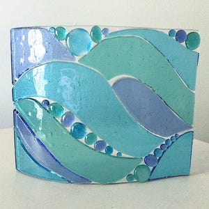 May include: A rectangular fused glass panel featuring a wave-like pattern in shades of turquoise, teal, and periwinkle blue. The design incorporates smooth, rounded glass pebbles of varying sizes scattered across the surface of the glass.