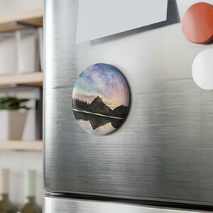 May include: A round magnet featuring a watercolor landscape of mountains reflected in a lake. The sky transitions from pink to blue, with a dark mountain silhouette and a forest line. The magnet is attached to a stainless steel refrigerator.