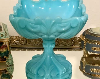 May include: A light blue milk glass compote dish featuring a square bowl with a scalloped rim and an ornate pedestal base. The glass has a smooth, opaque finish with molded decorative scrollwork patterns along the sides and the base.