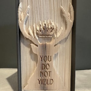 May include: A book folded into the shape of a stag with antlers. The stag is standing on a dark brown surface. The words "You Do Not Yield" are carved into the stag's body.