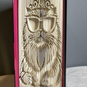 以下が含まれることがあります： サングラスと王冠をつけたフクロウのデザインが彫られた、ユニークなブックアート作品。 フクロウは本のページから彫られており、ピンクの背表紙と黒い縁があります。 アートワークは縦に表示されています。