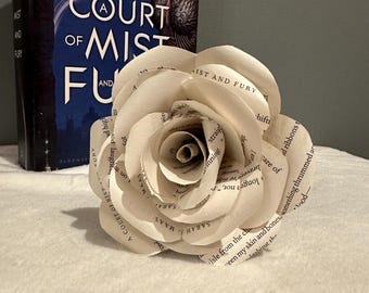 May include: A handmade paper rose made from book pages sits in front of a book titled "A Court of Mist and Fury" by Sarah J. Maas. The rose is white with visible text from the book pages. The book has a blue cover.