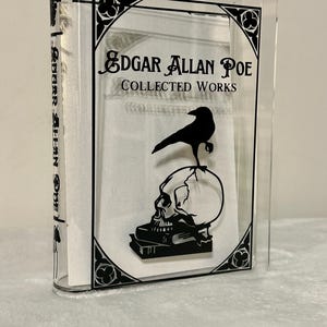 May include: A clear acrylic book display case with a black silhouette of a skull, a raven, and books. The text "Edgar Allan Poe Collected Works" is printed on the front of the case.