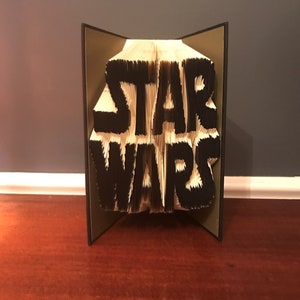 May include: A book folded into the shape of the "Star Wars" logo. The book is open and the logo is visible in black against a white background. The book is standing on a wooden surface.