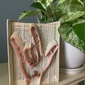 May include: A book art sculpture of a hand sign in a light pink color, made by folding the pages of a book. The sculpture is sitting on a wooden surface in front of a potted plant with green and white leaves.