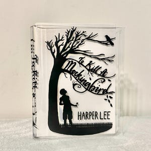 May include: A clear acrylic book cover with a black silhouette of a tree, a bird, a child, and the text "To Kill a Mockingbird" by Harper Lee.