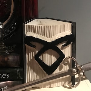 May include: A book with a black paper cover has been folded into the shape of a black symbol. The symbol is a stylized letter "X" with a circle around it. The book is sitting on a white surface with a silver sword lying on top of it.
