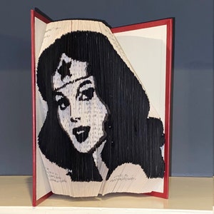 May include: A book art sculpture of Wonder Woman's face, created by folding the pages of a book. The sculpture is displayed on a white shelf against a dark blue wall.