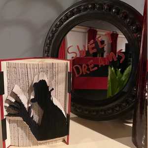 May include: A black silhouette of a person reading a book is folded from the pages of a book. The book is standing upright on a table. A black framed mirror with a red background is behind the book. The mirror has the words "Sweet Dreams" written on it in red.