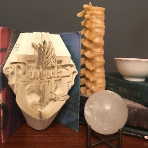 May include: A white papercraft sculpture of the Hogwarts crest, a book with a blue cover, a beige candle shaped like a spine, a clear crystal ball on a black stand, and a book with a black cover that says "Rogues" on the spine.