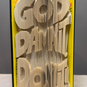 May include: A book sculpture featuring folded pages that form the words GOD DAMNIT DONUT. The book has a bright yellow cover with black edges visible on the sides. The paper edges are precisely folded to create the three-dimensional text effect.