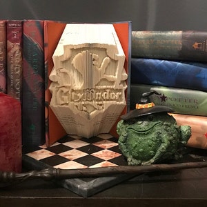 Op de afbeelding: Een boek gevouwen in de vorm van het Gryffindor-huiswapen uit de Harry Potter-serie. Het wapen is wit en heeft een leeuw. Het boek ligt op een zwart-wit geblokt oppervlak. Er staan ​​verschillende Harry Potter-boeken achter het boek.