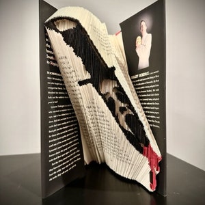 May include: A book with pages folded into the shape of a black and red knife. The book is open and the knife is sticking out of the pages. The back cover of the book has a black and white photo of a person holding a small object. The text on the back cover reads "Grady Hendrix is a master..."