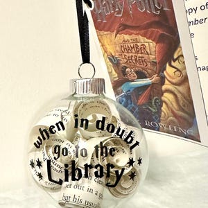 May include: Clear glass Christmas ornament filled with rolled paper and the text "when in doubt go to the Library" in black. The ornament hangs from a black ribbon. A book cover of "Harry Potter and the Chamber of Secrets" is in the background.