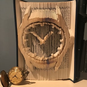 May include: A book art sculpture of a watch made by folding the pages of a book. The watch face is visible and the hands are pointing to the time. A small pocket watch is in the foreground.