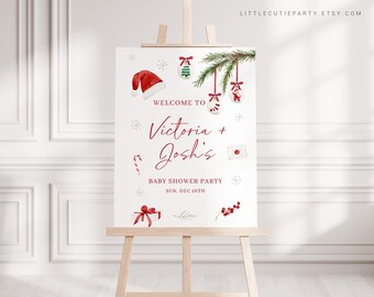 May include: A white baby shower party sign on a wooden easel. The sign has red text and illustrations of Christmas ornaments, a Santa hat, and a gift. The sign reads "Welcome to Victoria + Josh's Baby Shower Party Sun. Dec 26th."