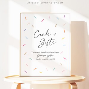 以下が含まれることがあります： カラフルなコンフェッティのデザインと、黒い筆記体で「Cards + Gifts」と書かれた白い看板。看板には、「Thank you for celebrating with us Jennifer Gates Sunday, Aug 26th - At 2 PM」というテキストも含まれています。