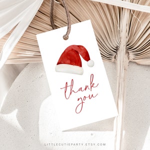 Peut inclure: Une &eacute;tiquette cadeau blanche avec une illustration &agrave; l'aquarelle d'un bonnet de P&egrave;re No&euml;l et l'inscription "thank you" en script rouge. L'&eacute;tiquette est suspendue par une ficelle marron, sur fond neutre.