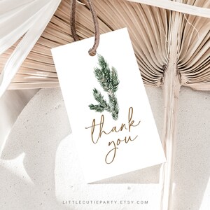 May include: A white rectangular gift tag with a watercolor illustration of green foliage and the words "thank you" in gold script. The tag is attached to a brown string and is set against a neutral background with dried palm leaves.