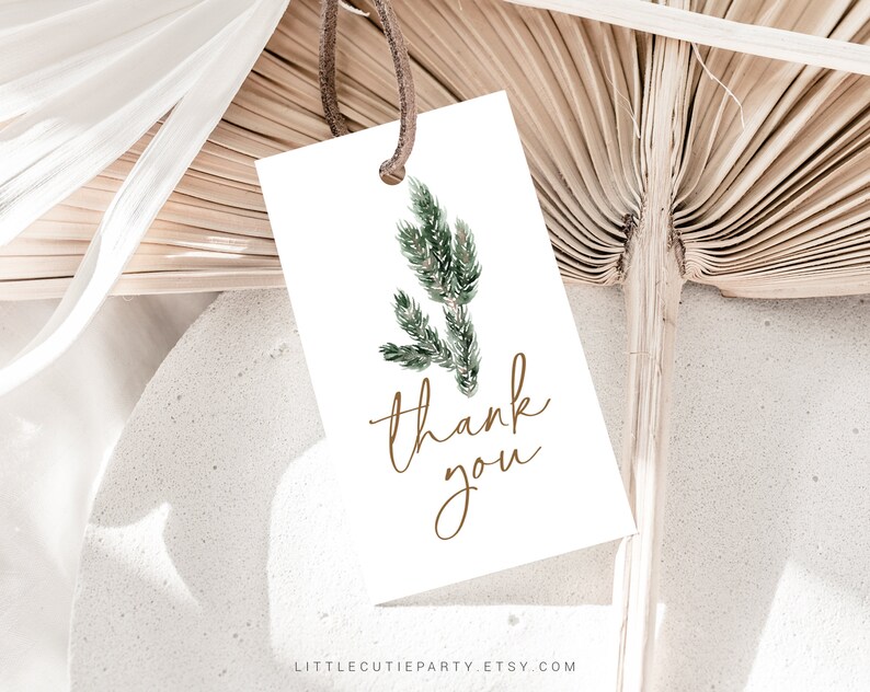 May include: A white rectangular gift tag with a watercolor illustration of green foliage and the words "thank you" in gold script. The tag is attached to a brown string and is set against a neutral background with dried palm leaves.