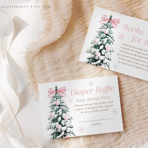 May include: Two baby shower cards with a watercolor Christmas tree design. One card reads "Books for Baby" and the other says "Diaper Raffle". Both cards have pink bows and snowflakes. The cards are on a cream-colored surface.