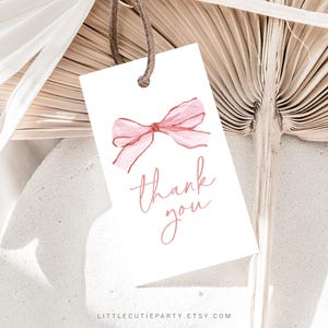 May include: A white gift tag with a watercolor-style pink bow and the words "thank you" in a delicate script. The tag is suspended by a brown string, perfect for adding a personal touch to gifts.