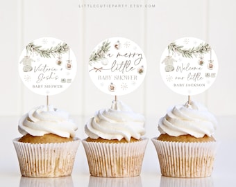 May include: Three cupcakes topped with white frosting and decorative toppers. The toppers feature watercolour illustrations of baby clothes, foliage, and text saying "Victoria + Josh's BABY SHOWER", "a merry little BABY SHOWER", and "Welcome our little BABY JACKSON".