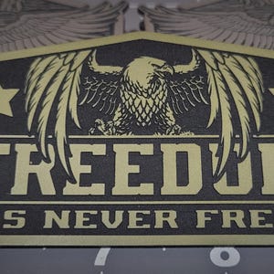 May include: A rectangular patch with a black background and gold lettering and design. The patch features an eagle with outstretched wings, two stars, and the words "FREEDOM IS NEVER FREE."